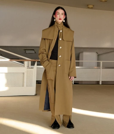 A full-length shot captures a singular adult female showcasing a tan trench coat with statement buttons and matching pants in a neutral setting.