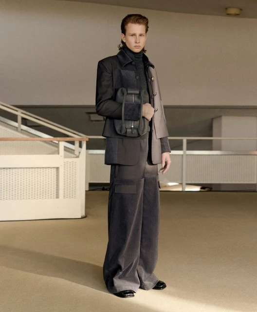 A young adult man poses indoors wearing a grey jacket, turtleneck, wide-legged pants, and carrying a small black bag.