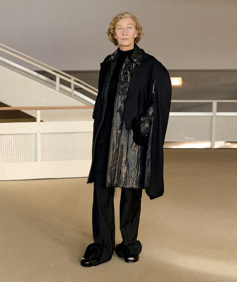 Elderly woman standing in a modern building interior wearing a layered black coat and trousers for William Fan AW25 lookbook.