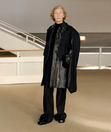 Elderly woman standing in a modern building interior wearing a layered black coat and trousers for William Fan AW25 lookbook.
