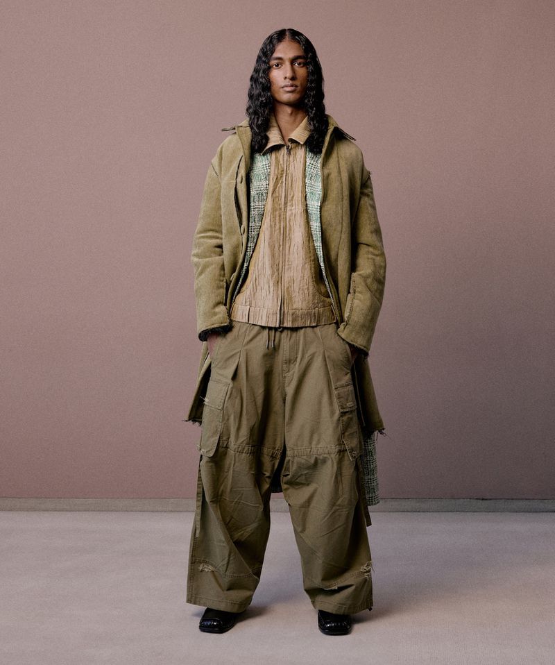 Full-length fashion shot: a male model with long hair wearing a neutral coat, shirt, and baggy pants, standing against a solid tan backdrop.