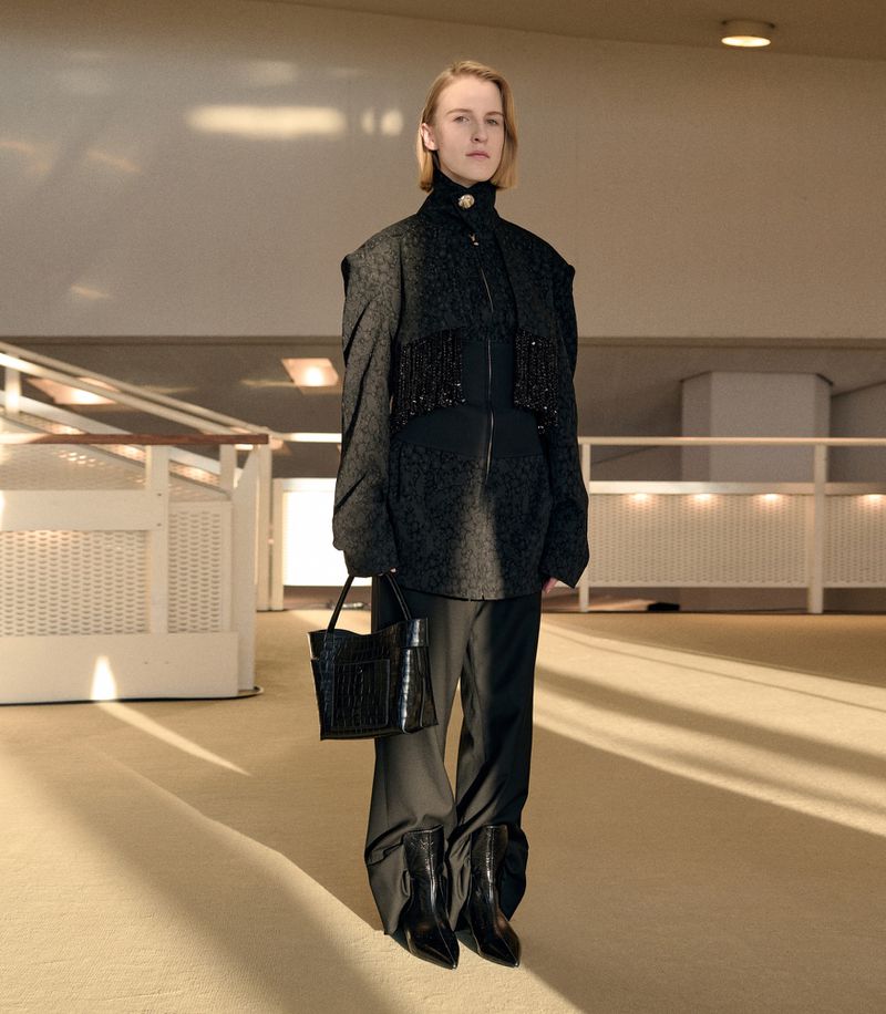 Model wearing dark textured jacket, trousers, and holding handbag inside a modern architectural interior by Léa Wormsbach.