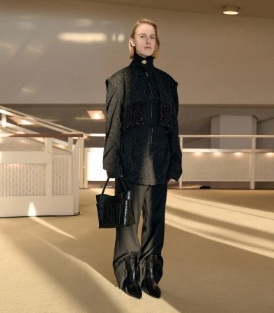 Model wearing dark textured jacket, trousers, and holding handbag inside a modern architectural interior by Léa Wormsbach.