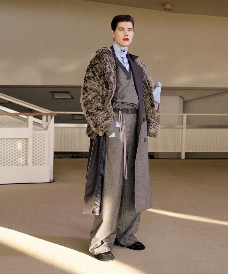 Fashionable adult in a fur-collared coat, grey suit, and a light blue shirt. Indoor setting.