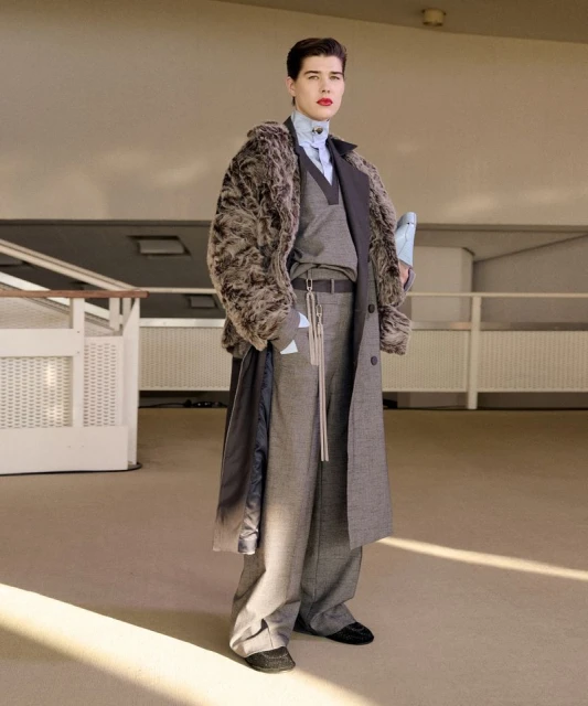 Fashionable adult in a fur-collared coat, grey suit, and a light blue shirt. Indoor setting.