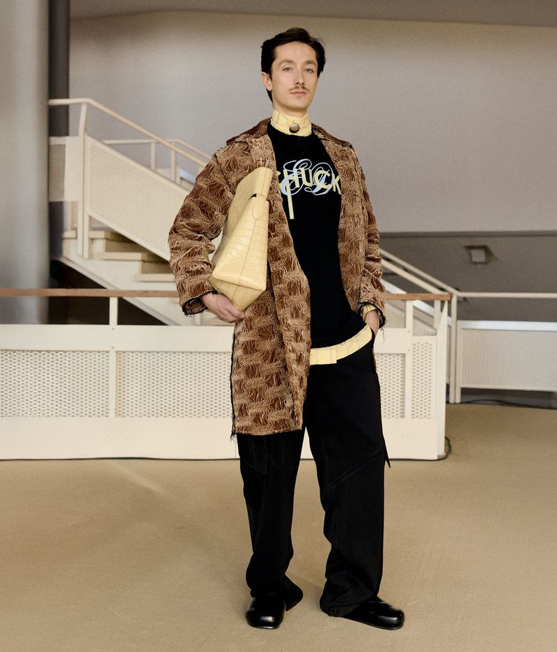 An adult male stands indoors, wearing a brown patterned coat over a black shirt, baggy black pants, and black shoes, holding a cream bag.