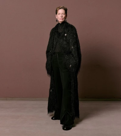Model standing against a plain brown background wearing a textured black long coat and dark trousers for William Fan AW25 lookbook.
