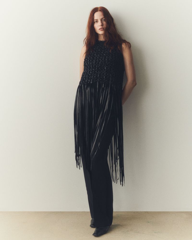 A red-haired woman in a sleeveless black fringe dress and wide-leg pants poses indoors against a neutral wall in a full-length shot.