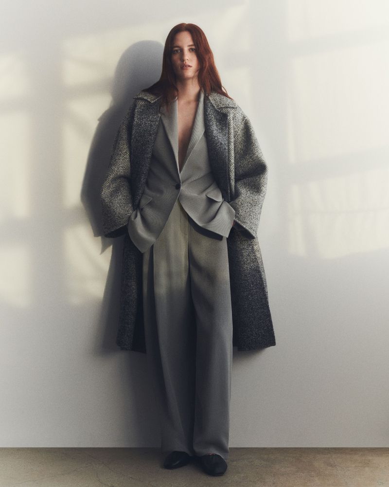 Fashion shot of a red-haired woman posing in an oversized grey suit and coat in front of a textured white wall with sunlight.