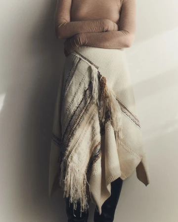 Model wearing a neutral long-sleeve top and an asymmetric cream knit skirt with fringed striped detailing against a plain wall.