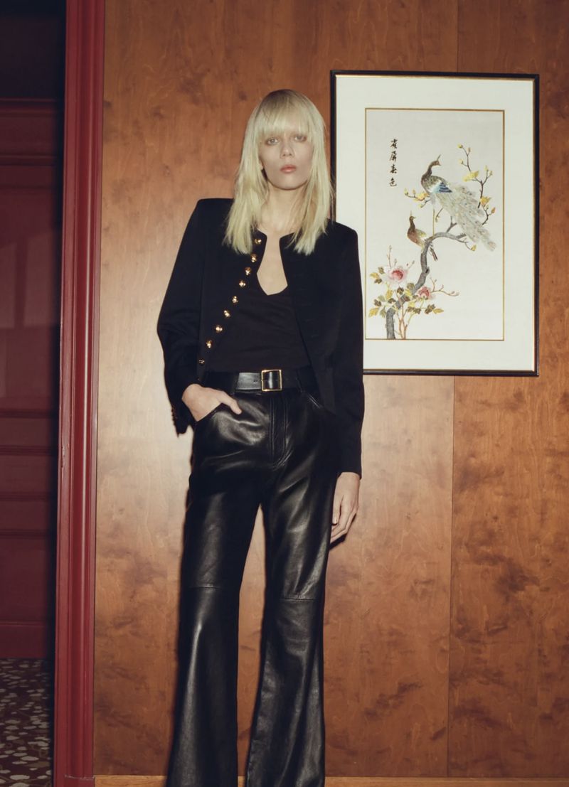 A woman in a black blazer and leather pants stands indoors in a fashion shot. She has blonde hair and fair skin, posing near a wood panel.