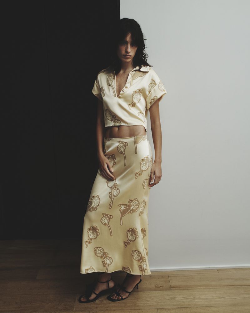 Woman in pale yellow skirt and blouse with cartoon pattern posing against black and white wall in indoor fashion shot.