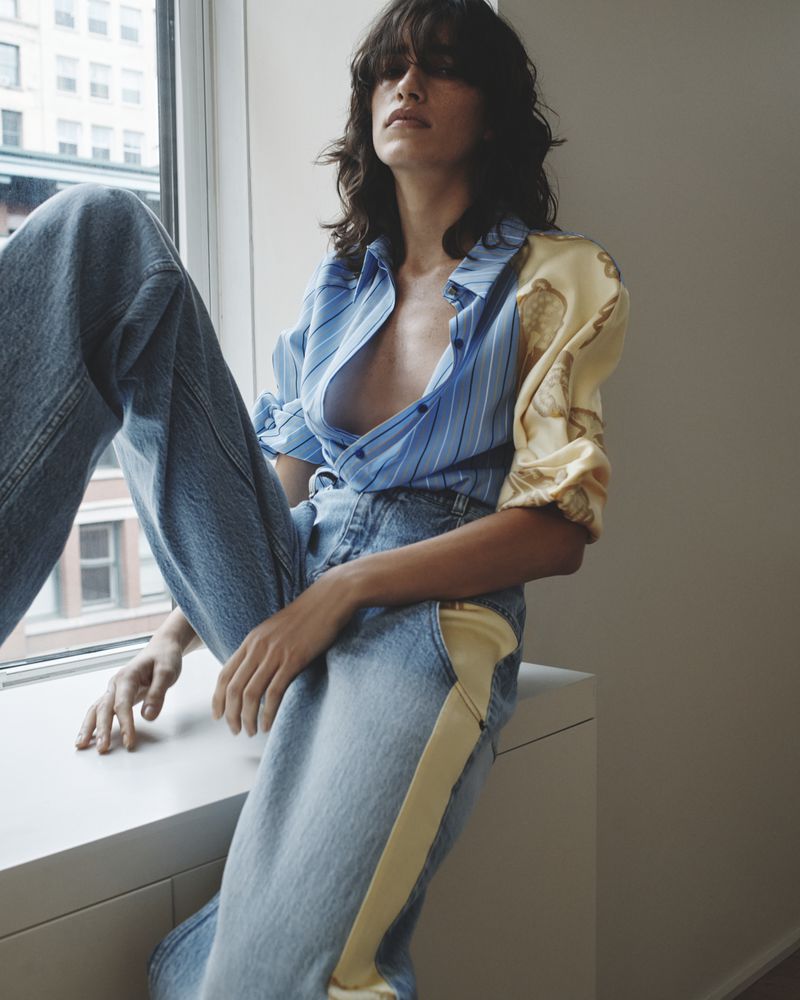 Young woman in unbuttoned blue striped shirt and baggy jeans sitting in window light, posing for fashion editorial photography.