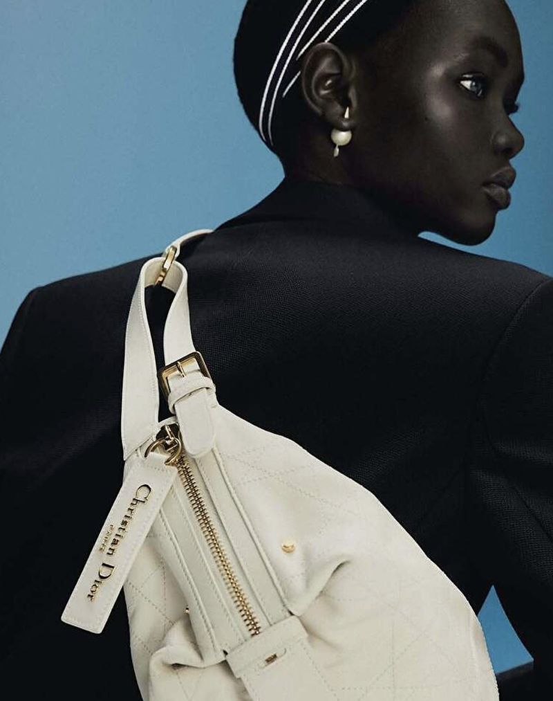 Close-up studio shot of a dark-skinned adult woman wearing a black jacket, pearl earrings and a cream-colored handbag with the brand logo.