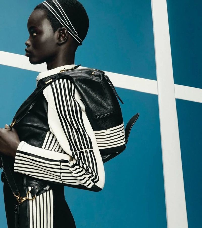 Dark-skinned woman with black-and-white-striped top, black backpack, and striped headband poses against a blue-and-white background.