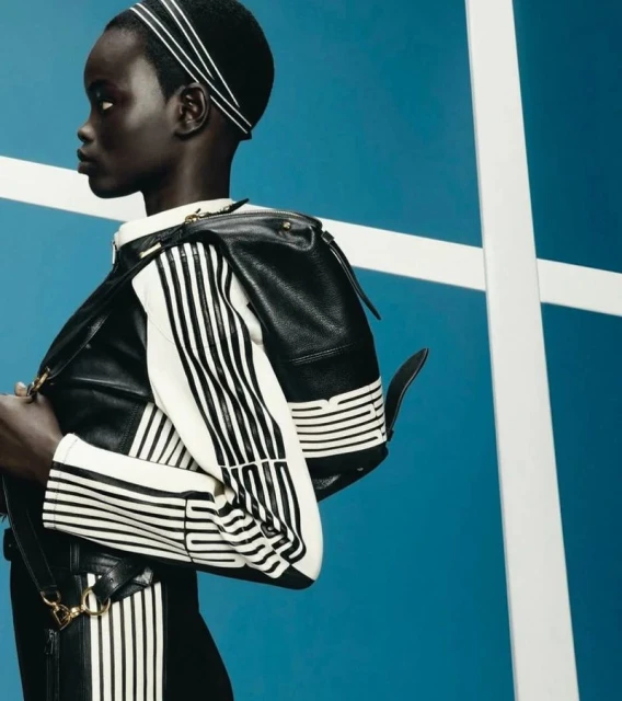 Dark-skinned woman with black-and-white-striped top, black backpack, and striped headband poses against a blue-and-white background.