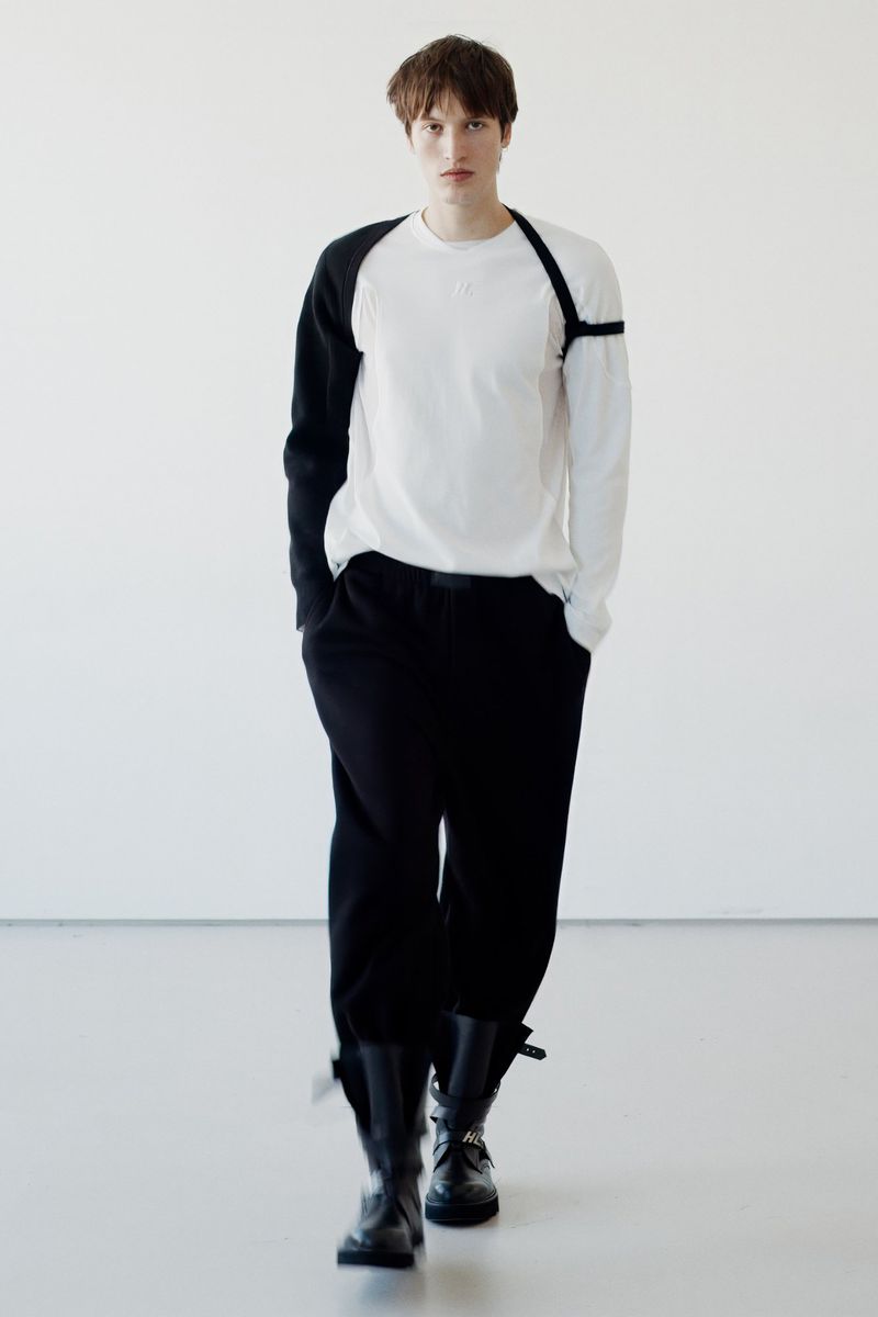 Male fashion model walks toward the camera in a bright studio wearing black boots, black pants, and a two-tone white-and-black long-sleeved shirt.
