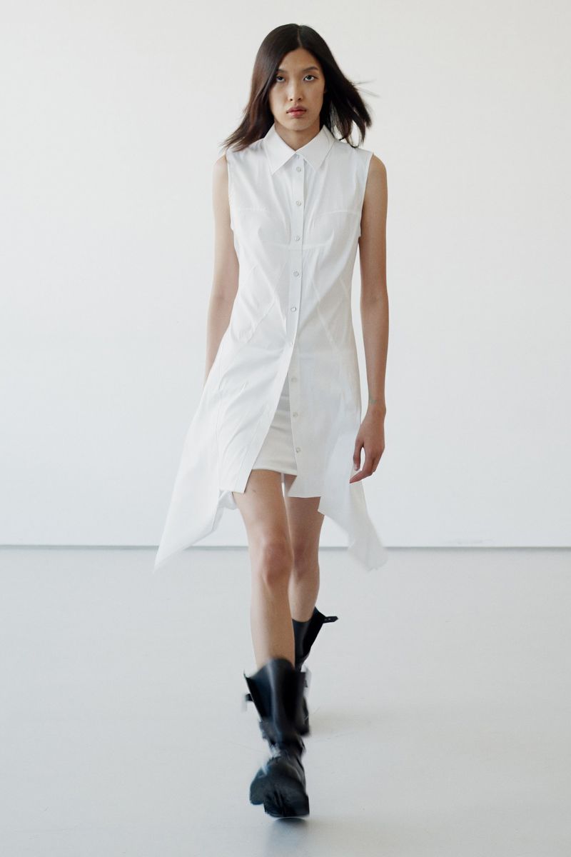 Model walking forward wearing a sleeveless white button-down dress and black boots against a plain white studio backdrop.