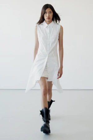 Model walking forward wearing a sleeveless white button-down dress and black boots against a plain white studio backdrop.