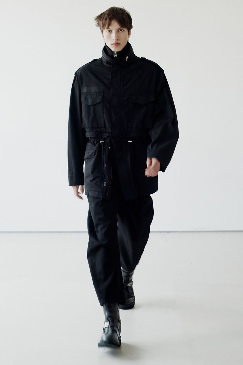 Young adult strides forward in a black utility jacket, pants, and buckle boots against a neutral backdrop in this full-length fashion shot.