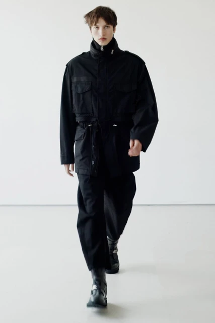 Young adult strides forward in a black utility jacket, pants, and buckle boots against a neutral backdrop in this full-length fashion shot.
