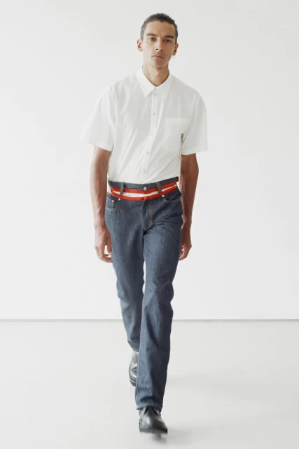 An adult male model in a white shirt and blue jeans walks in a studio setting, showcasing fashion and style with a confident expression.