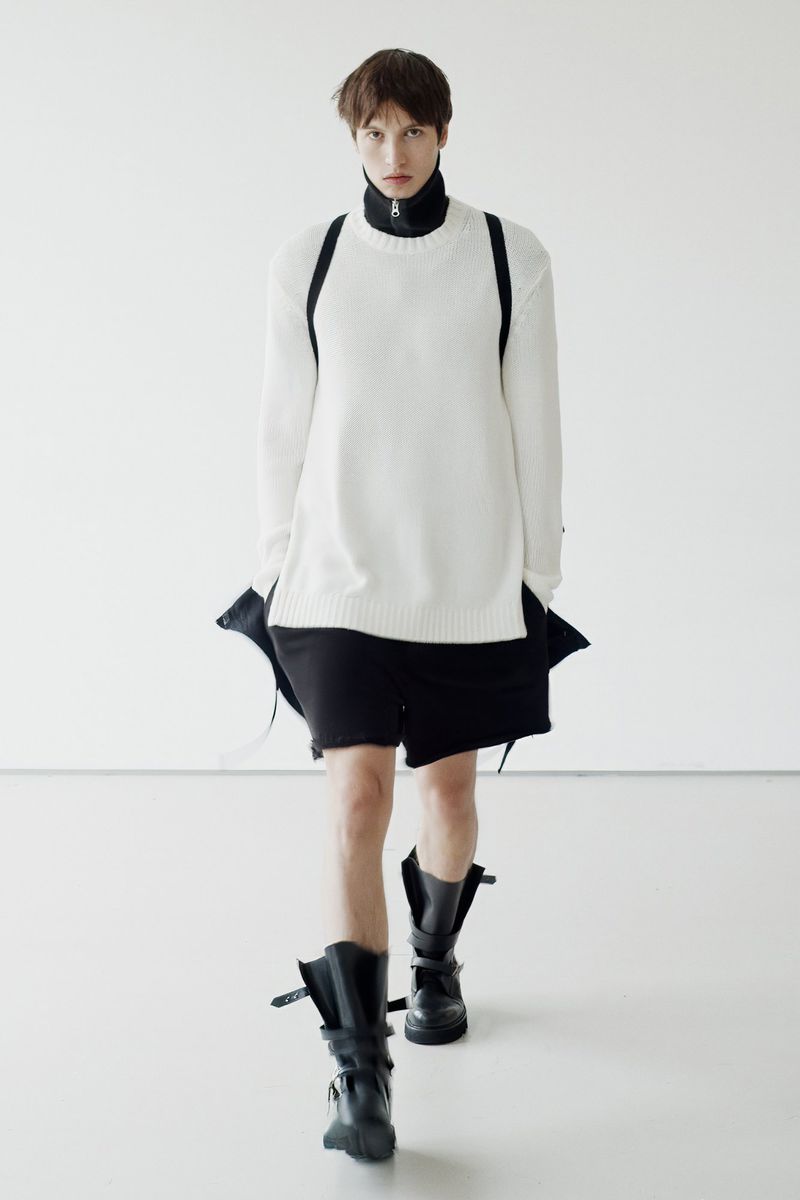 Fashion model in a white sweater and black shorts walks in a brightly lit photo studio wearing black platform boots with buckles.