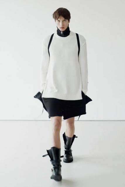 Fashion model in a white sweater and black shorts walks in a brightly lit photo studio wearing black platform boots with buckles.