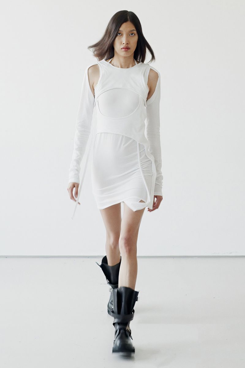 Fashion model in a sleeveless white dress and black boots walks in a minimalist white studio with bright, soft lighting. 