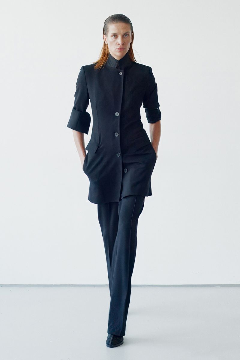A woman poses in a black, button-up outfit and pants against a bright white background. The image focuses on fashion and style.