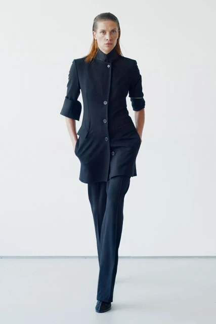 A woman poses in a black, button-up outfit and pants against a bright white background. The image focuses on fashion and style.