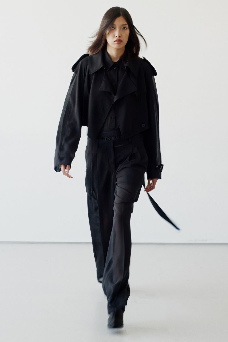 A runway shot showcases a model in an all-black outfit, including a cropped jacket and wide-leg pants, walking against a plain white background.
