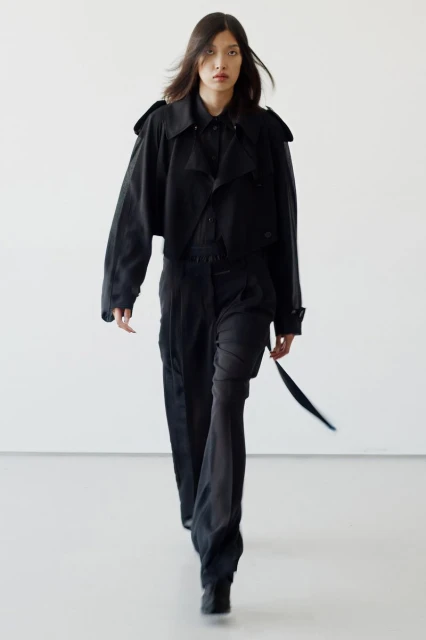 A runway shot showcases a model in an all-black outfit, including a cropped jacket and wide-leg pants, walking against a plain white background.