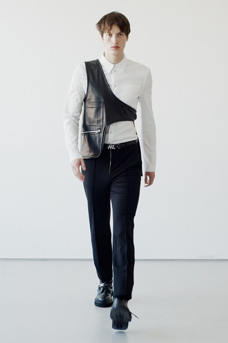 Male fashion model walks forward wearing white shirt, black vest, and black pants against a stark white background in a studio setting.