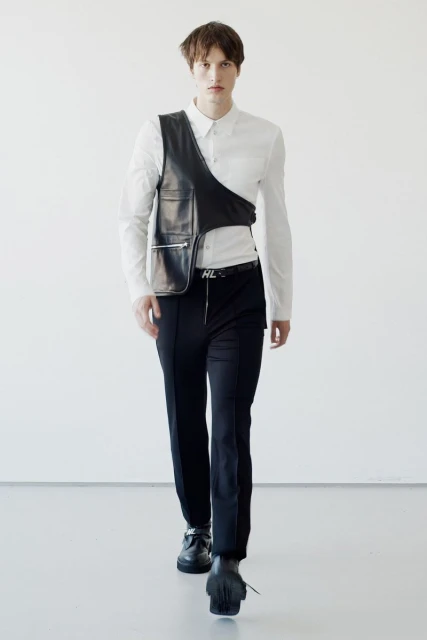 Male fashion model walks forward wearing white shirt, black vest, and black pants against a stark white background in a studio setting.