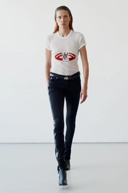 A woman in a white tee and black pants walks towards the camera in a stark white setting. Modern fashion, studio lighting, minimalist design.