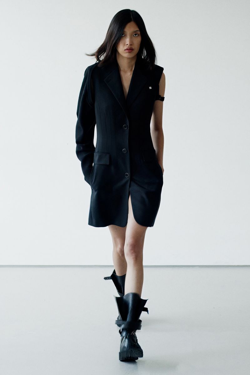 Runway fashion: An Asian woman strides in a black blazer dress, accented by sleek, dark hair, set against a crisp white backdrop.