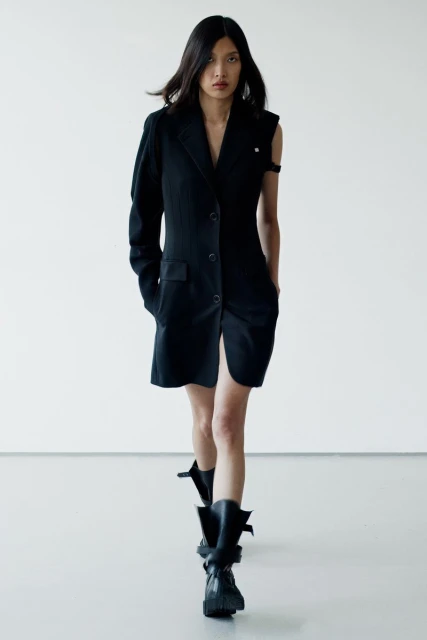 Runway fashion: An Asian woman strides in a black blazer dress, accented by sleek, dark hair, set against a crisp white backdrop.