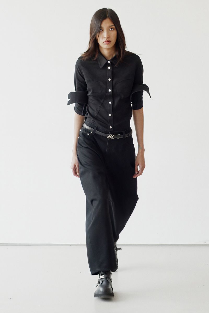 An adult female fashion model in a black shirt and wide-leg black jeans walking in a photo studio with a plain white background.