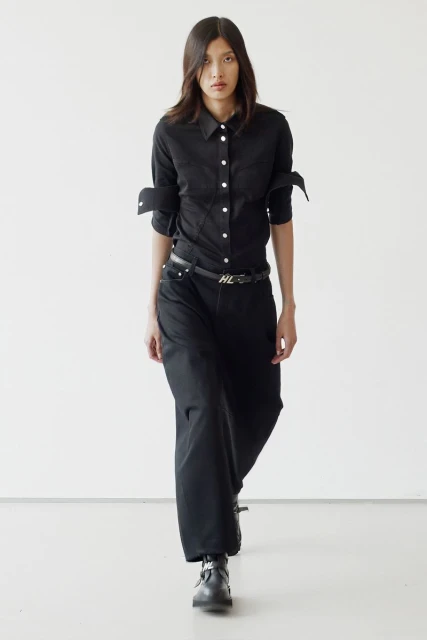 An adult female fashion model in a black shirt and wide-leg black jeans walking in a photo studio with a plain white background.