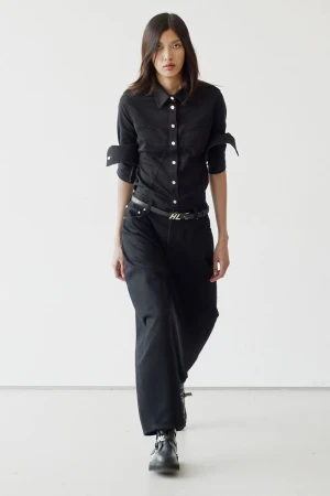 An adult female fashion model in a black shirt and wide-leg black jeans walking in a photo studio with a plain white background.