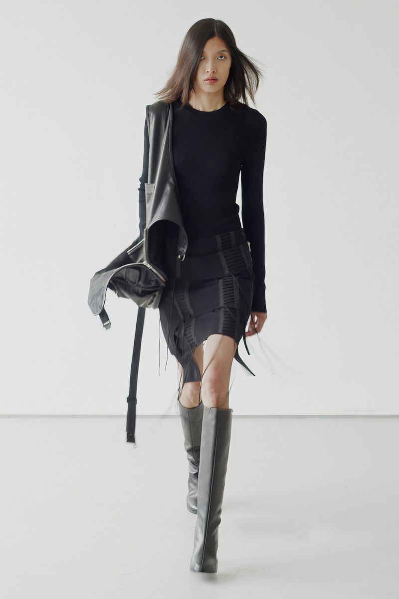 A person in dark clothing walks toward the camera in a studio. She wears a black top, black skirt, gray boots, and a black leather satchel.