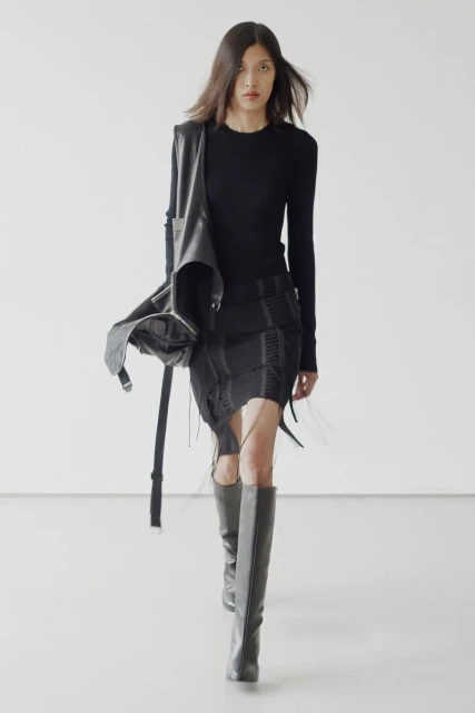 A person in dark clothing walks toward the camera in a studio. She wears a black top, black skirt, gray boots, and a black leather satchel.