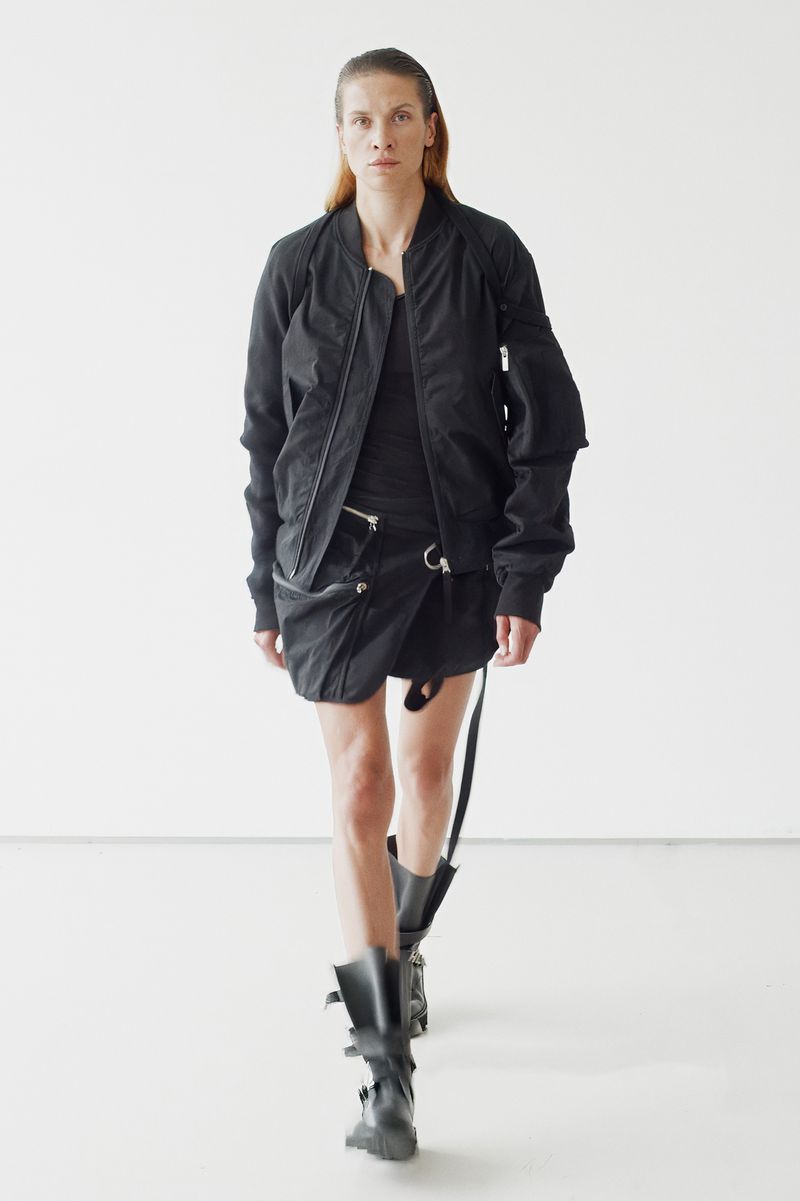 Runway model in black bomber jacket, dark top, wrap skirt, and buckled boots walks against white backdrop in studio lighting for fashion shoot.