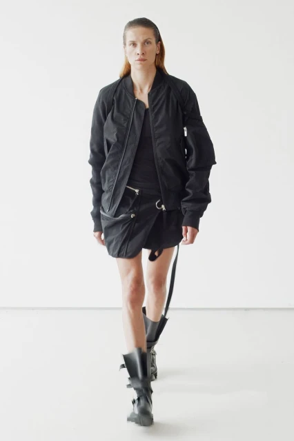 Runway model in black bomber jacket, dark top, wrap skirt, and buckled boots walks against white backdrop in studio lighting for fashion shoot.