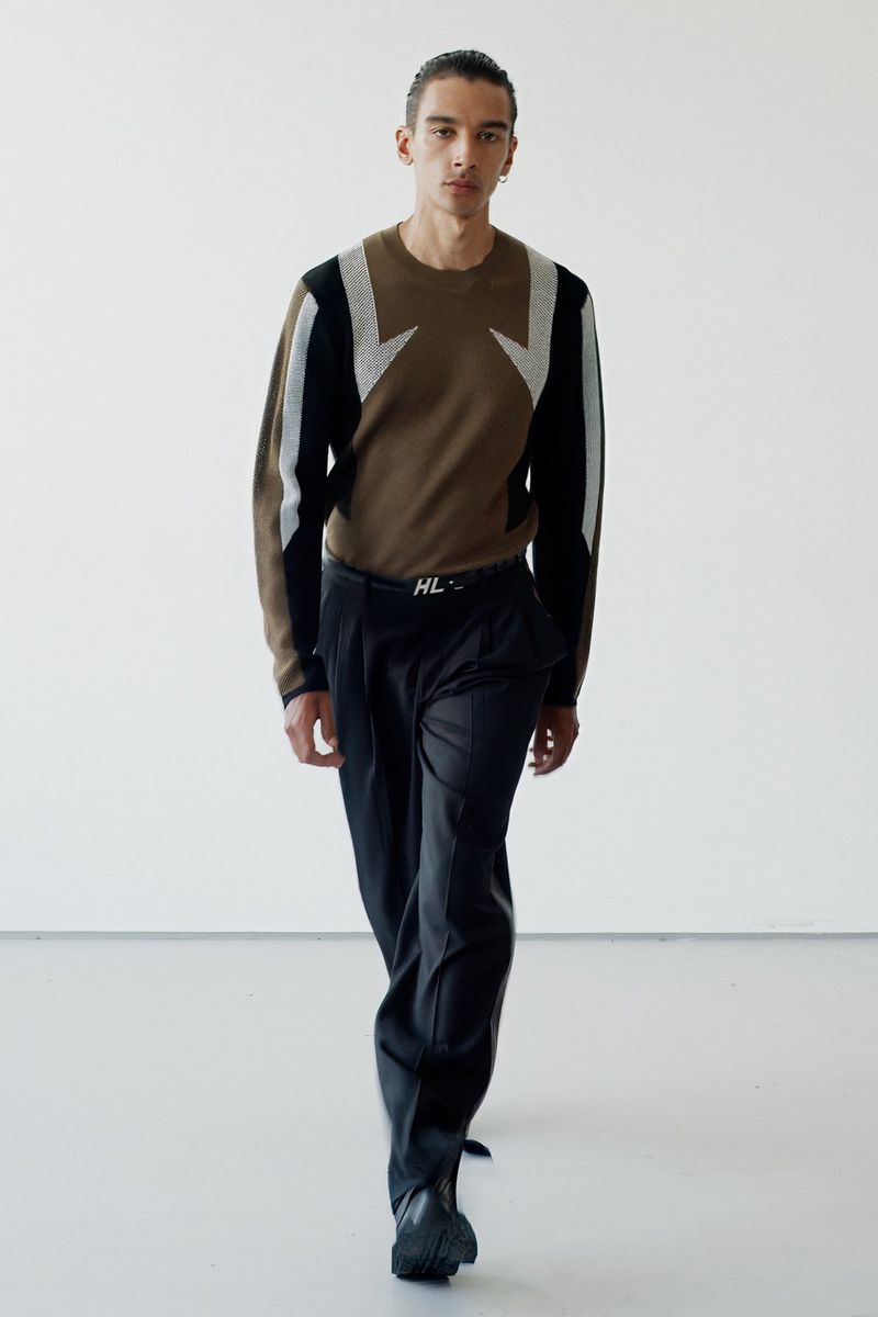 Full-length shot of a male model in a brown graphic sweater and black trousers against a white studio backdrop, fashion editorial.