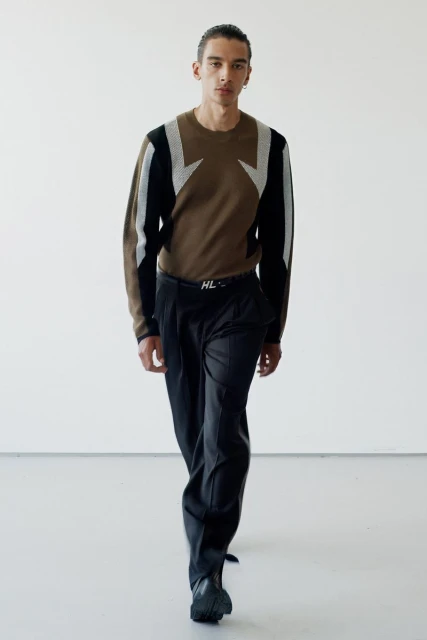Full-length shot of a male model in a brown graphic sweater and black trousers against a white studio backdrop, fashion editorial.