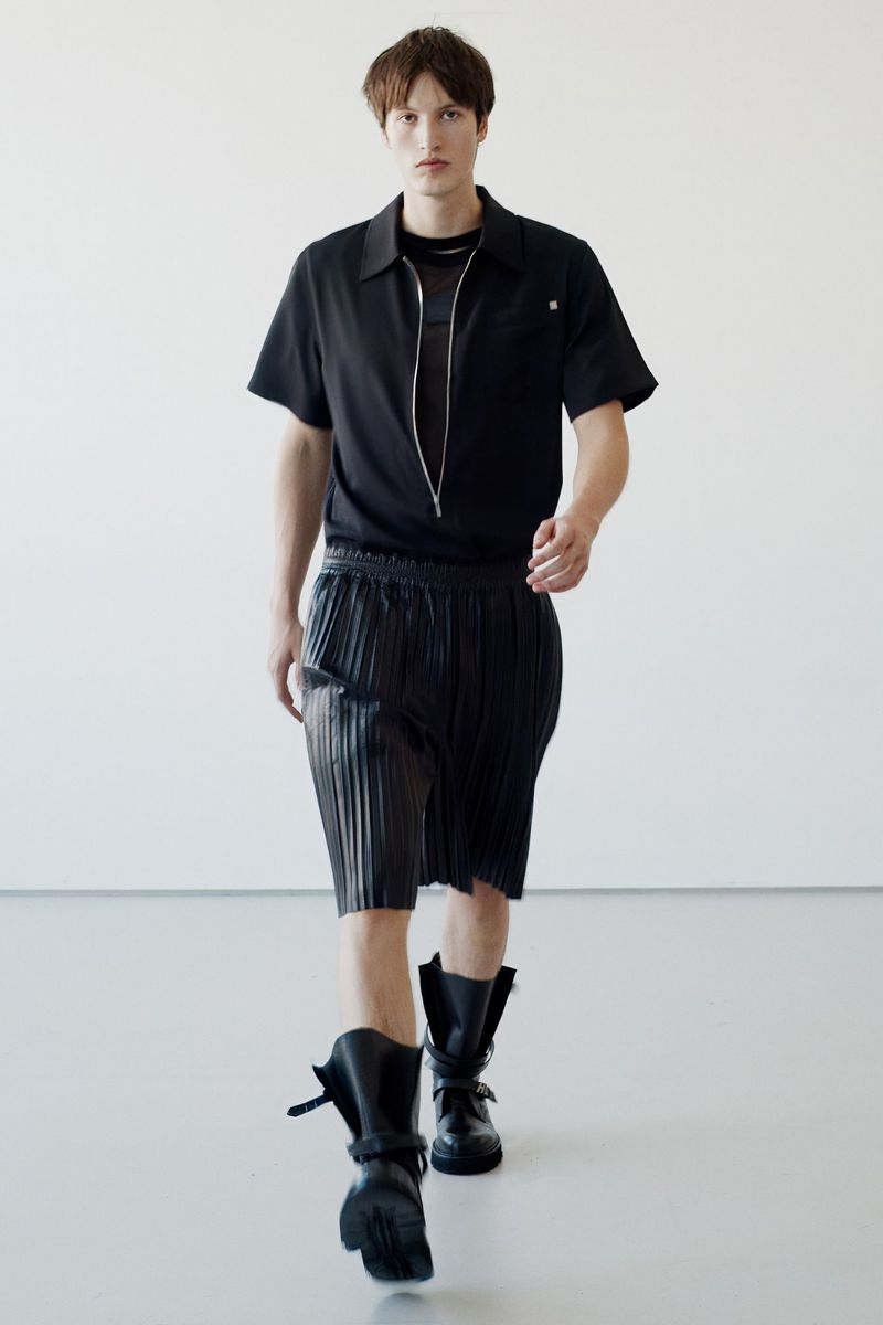 Male fashion model walks in black short-sleeved shirt, pleated shorts, and black lace-up boots against a white backdrop.