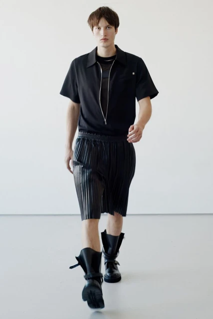 Male fashion model walks in black short-sleeved shirt, pleated shorts, and black lace-up boots against a white backdrop.