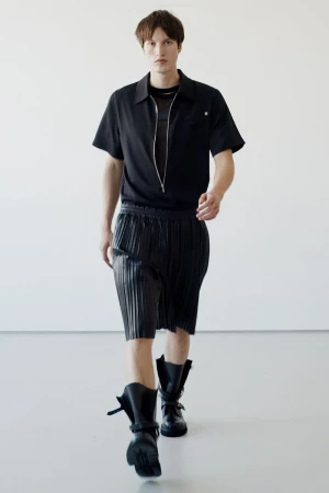 Male fashion model walks in black short-sleeved shirt, pleated shorts, and black lace-up boots against a white backdrop.